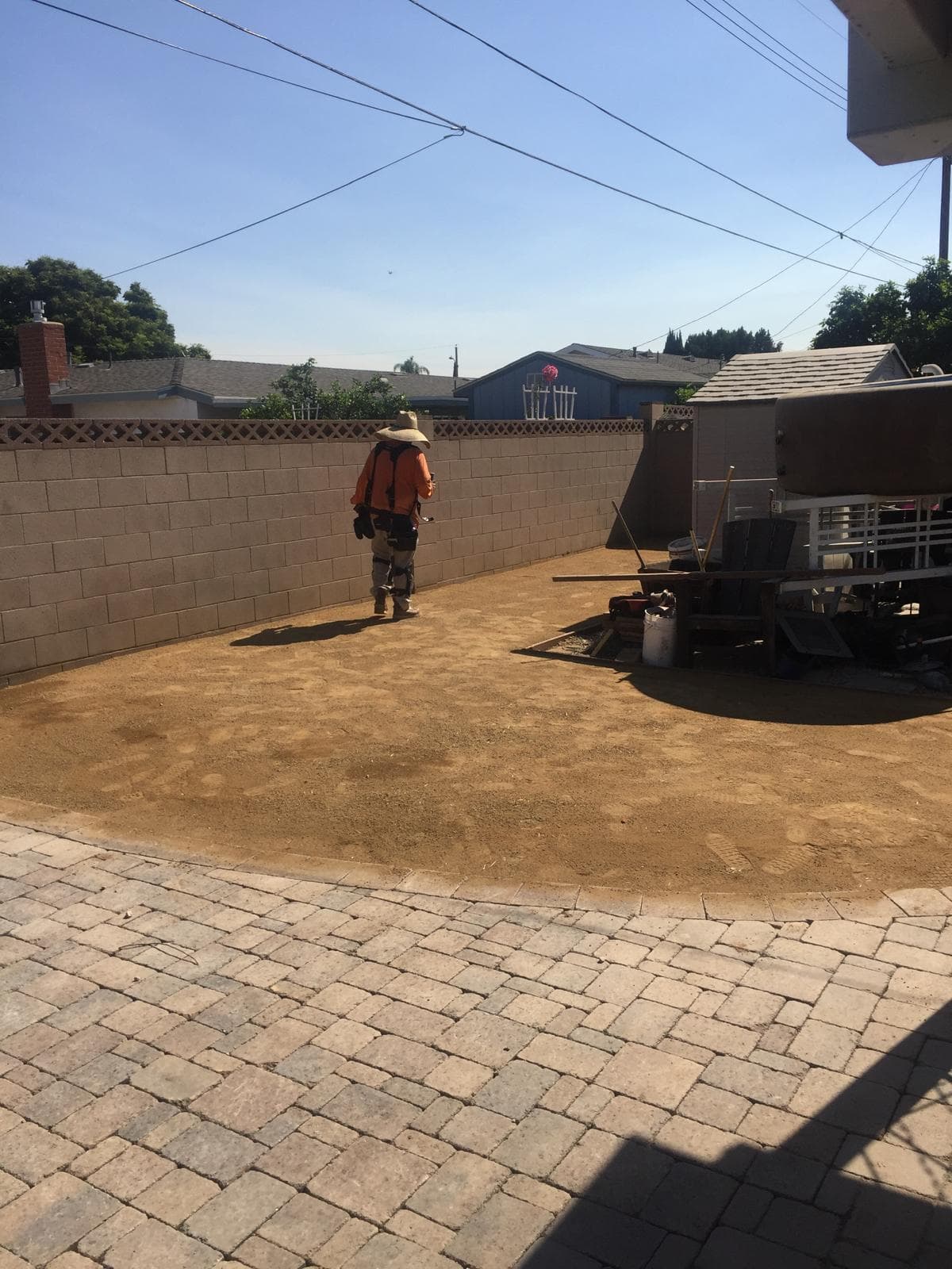A worker wearing a sun hat walks across a dirt area prepared for backyard landscaping.