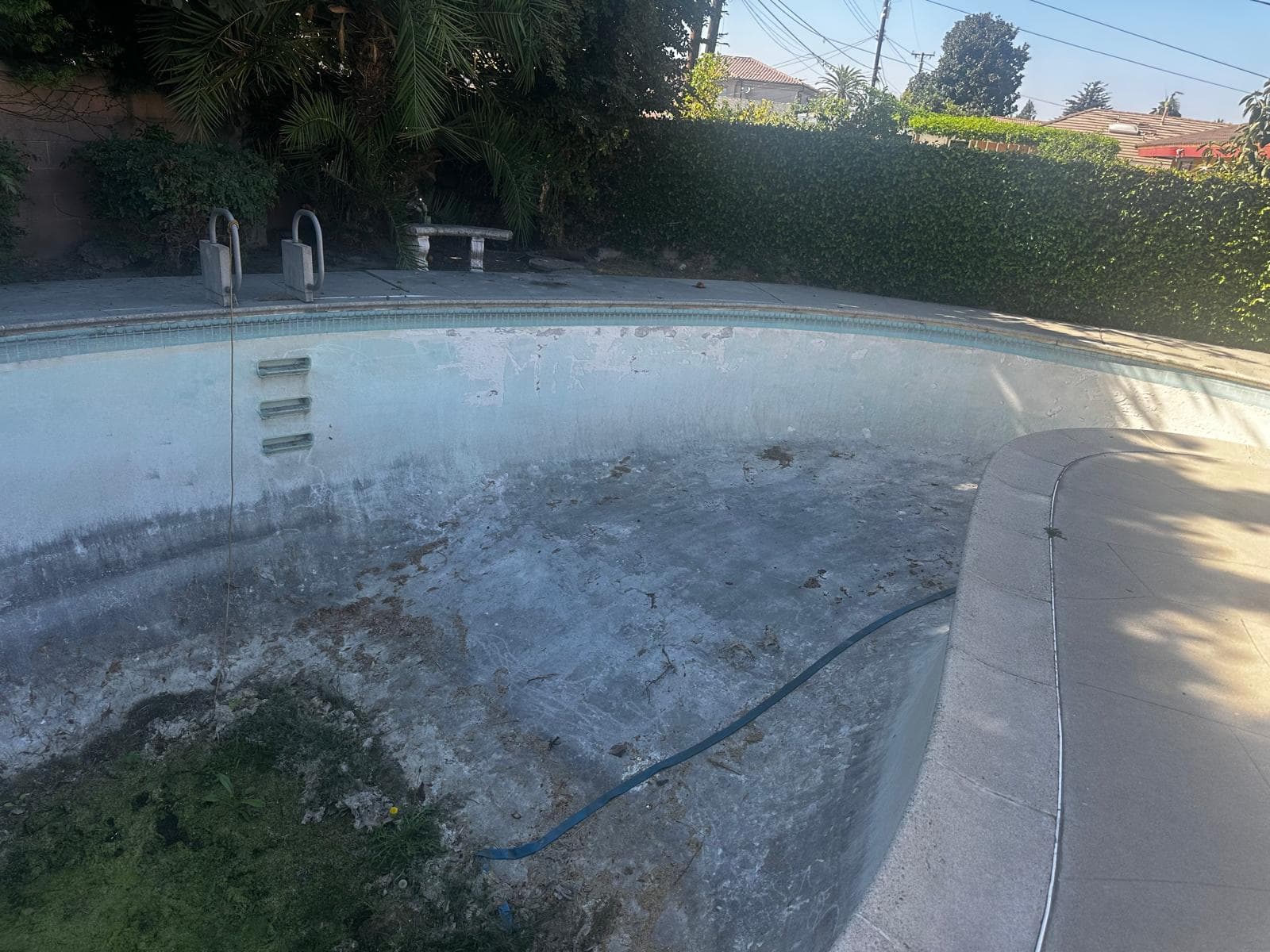 Empty, neglected swimming pool with stained walls and debris in a sunny residential backyard.